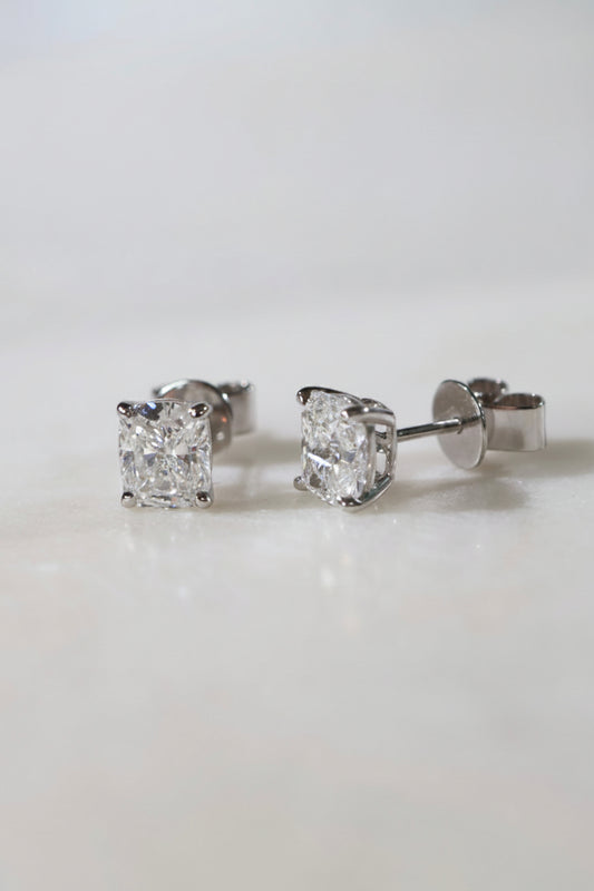 A pair of elongated cushion-cut diamond stud earrings in silver four-prong settings, shown on a clean white surface with a soft-focus background.